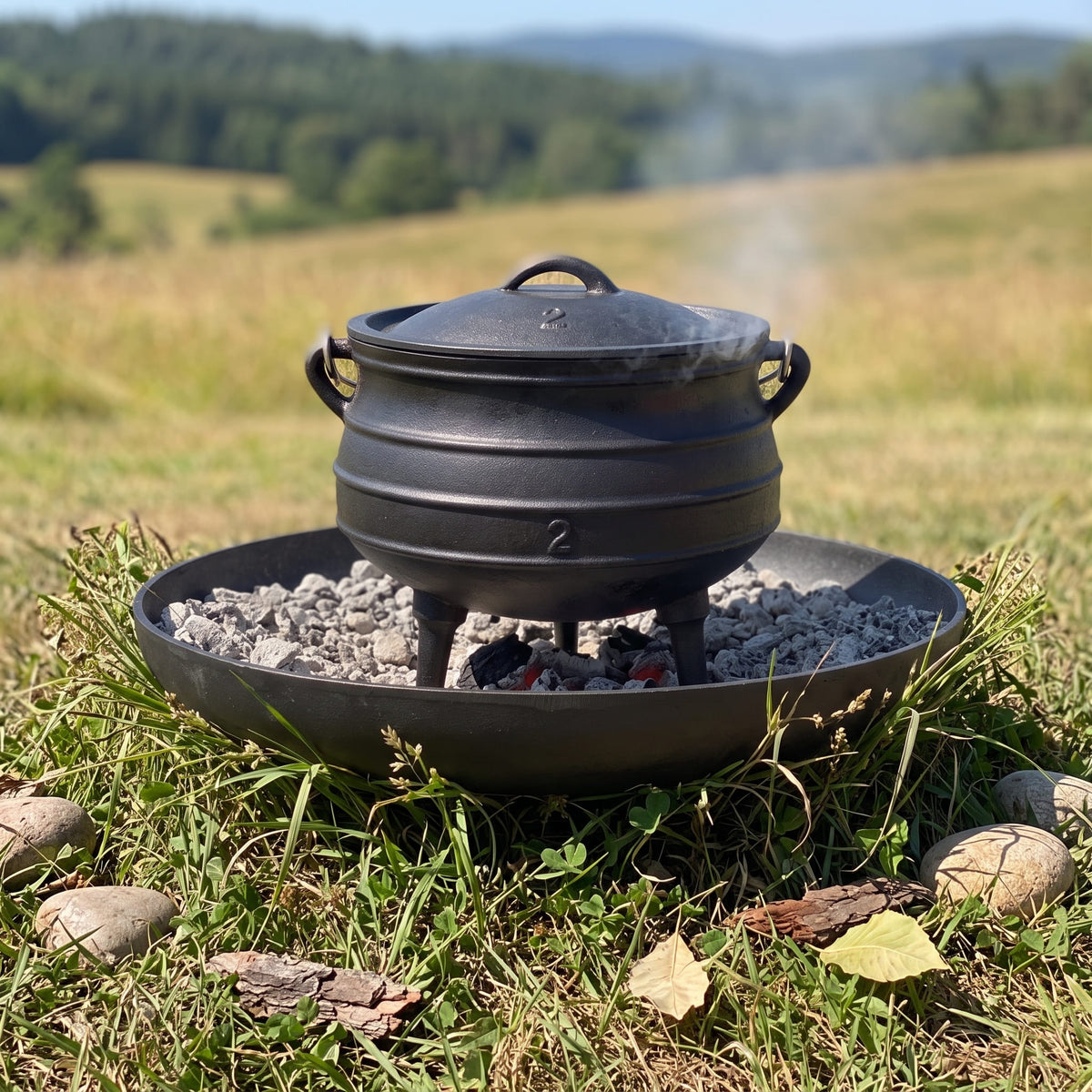 A black cast iron Dutch oven with a lid sits over a round metal fire pan containing coals and gray stones, placed outdoors in grassy terrain with a blurred field and trees in the background. 6 qt (Size #2), 8 qt (Size #3), 10 qt (Size #4), 14 qt (Size #6)