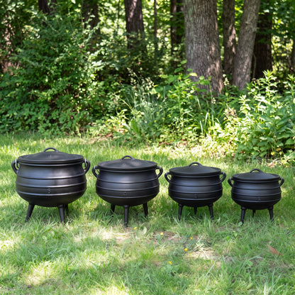 Four black cast-iron cauldrons of decreasing sizes arranged on grass in a wooded outdoor setting with trees and greenery in the background. 6 qt (Size #2), 8 qt (Size #3), 10 qt (Size #4), 14 qt (Size #6)