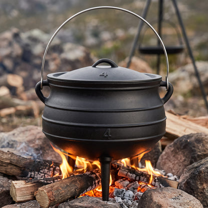 A black cast iron Dutch oven pot with a lid and metal handle, shown outdoors over an open fire with logs and rocks in a natural outdoor setting. 10 qt (Size #4) cast iron potjie