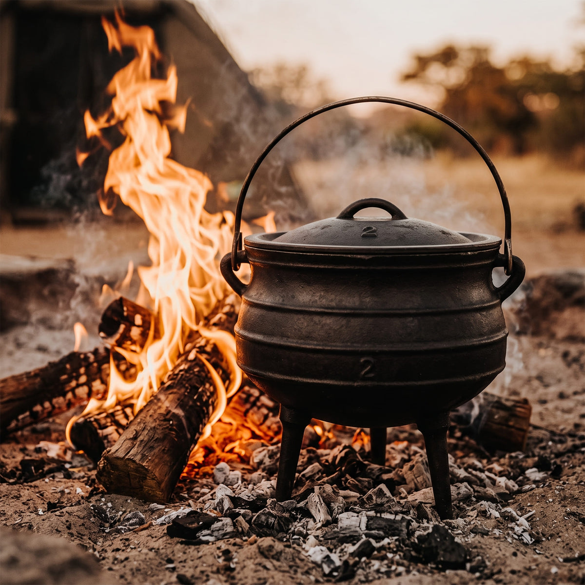 A black cast iron cauldron with a lid and triple legs sits over an open campfire with bright orange flames and burning logs, set outdoors against a softly blurred background. 6 qt (Size #2) cast iron potjie