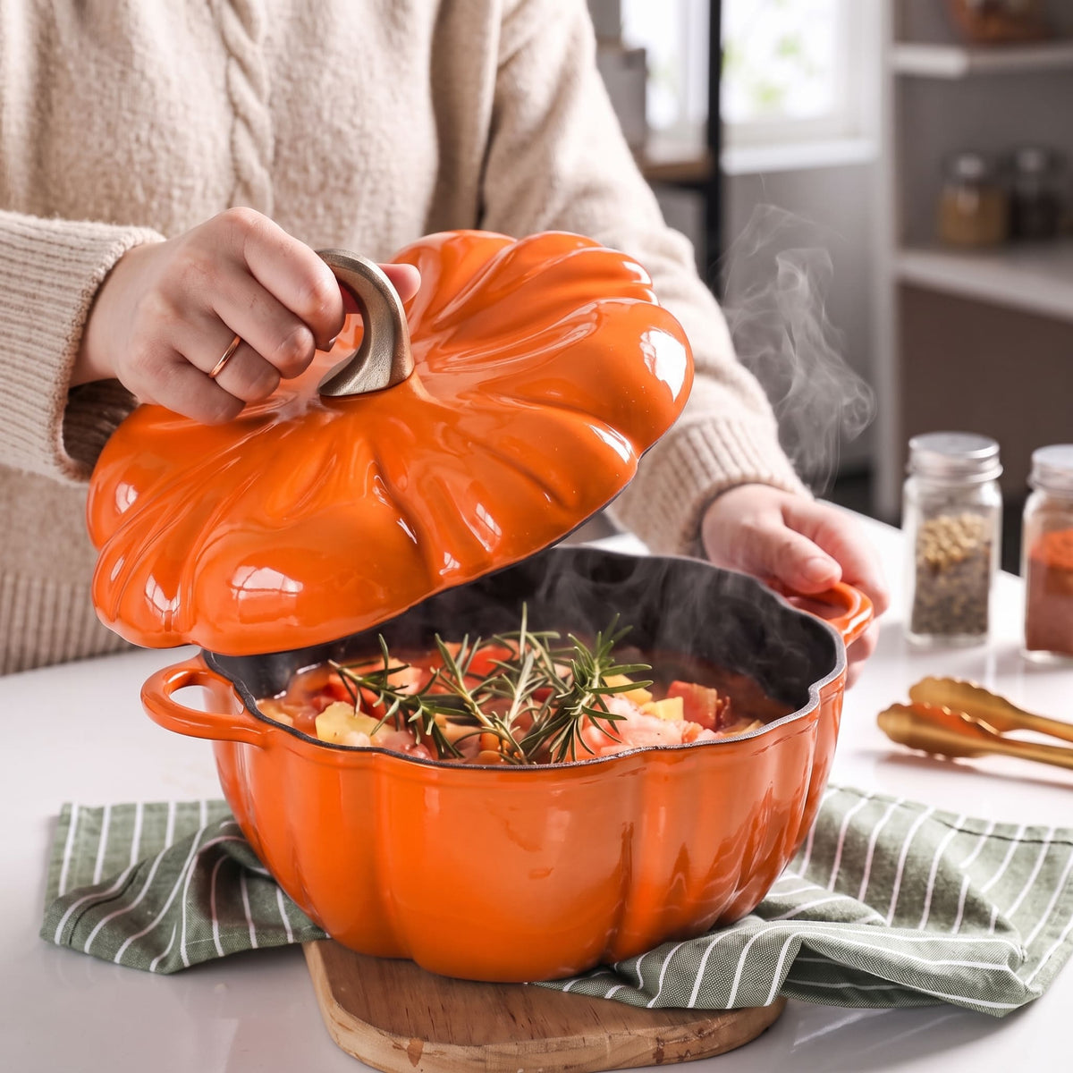 An pumpkin orange pumpkin-shaped enameled cast iron Dutch oven with a matching lid, being lifted by a person in a cozy kitchen setting. The pot is filled with a steaming autumn stew containing vegetables and rosemary, resting on a green striped cloth.