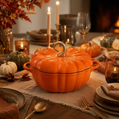 An orange enameled lidded casserole dish with a sculpted pumpkin handle and loop lid, styled on a rustic table runner with candles, plates, and autumn decorations.