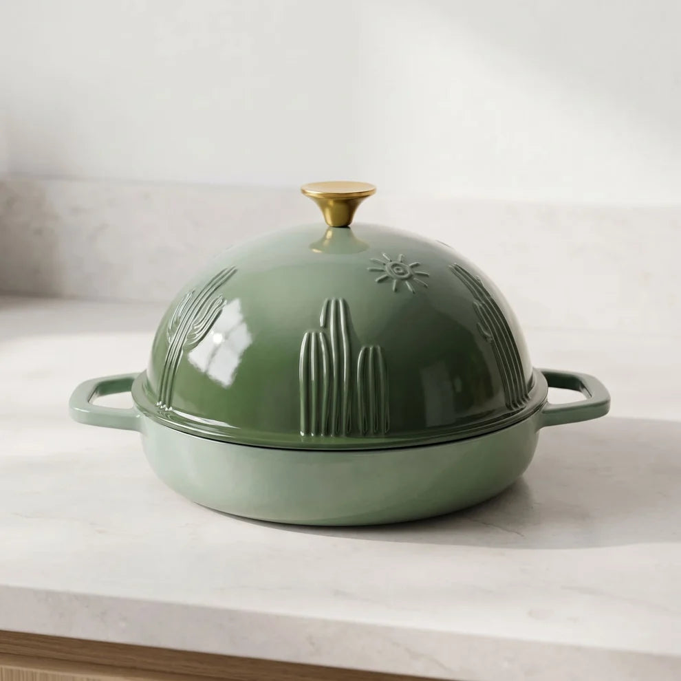 A sage green cast iron bread oven with a matching cloche lid, featuring embossed desert cactus and sun motifs, resting on a light marble countertop.
