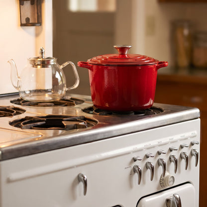 A cherry red enameled cast iron Dutch oven with a lid sits on a white retro-style gas stove in a warm kitchen setting, next to a clear glass teapot.