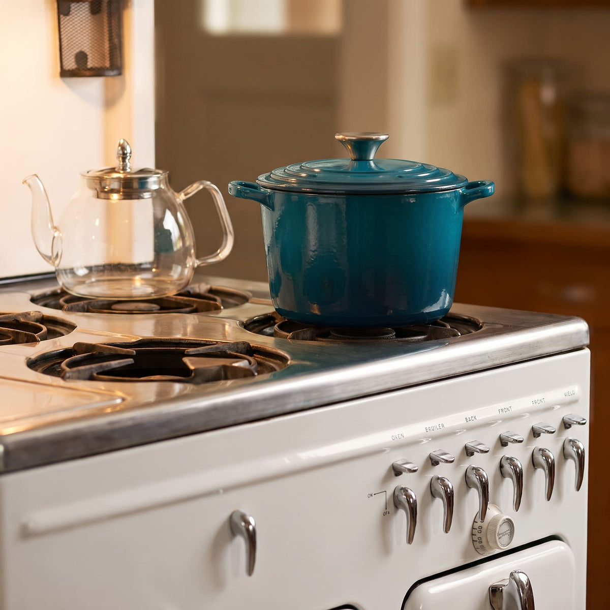 A deep cyan enameled cast iron Dutch oven with a lid sits on a white vintage-style gas stove, alongside a clear glass teapot.