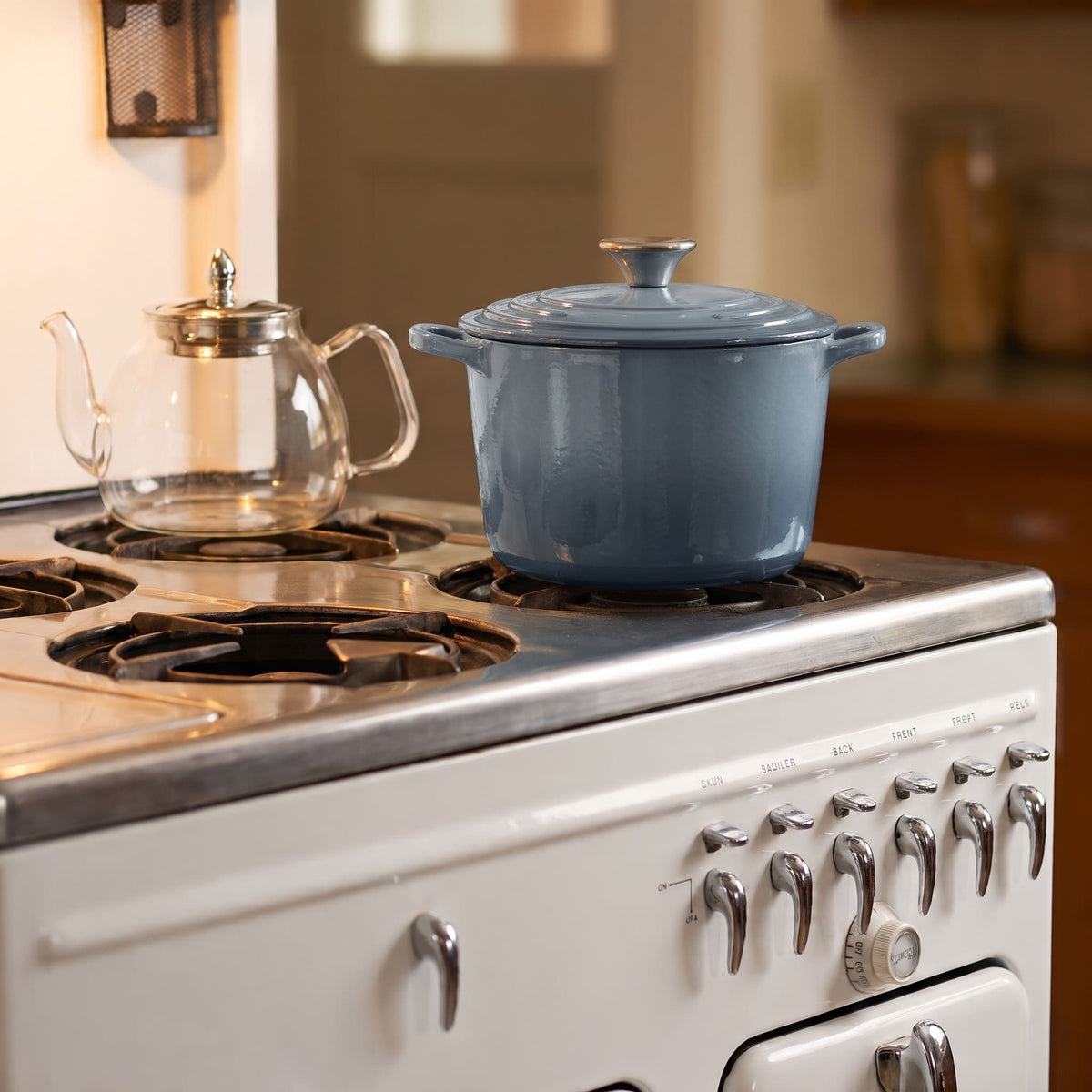 A slate blue enameled cast iron Dutch oven with a lid sits on a white vintage-style gas stove, with a clear glass teapot on a burner in a warm kitchen setting.