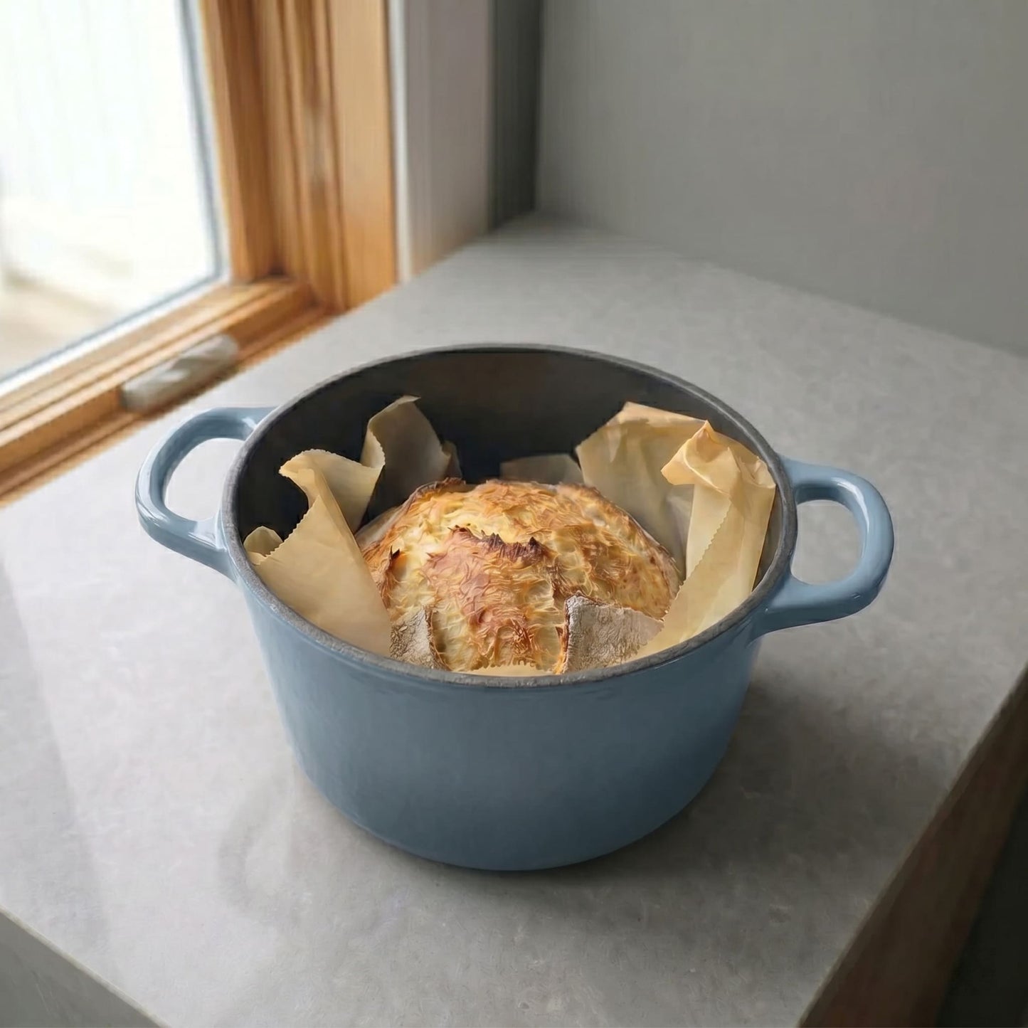 Blue deep Dutch oven holding a round loaf in parchment with side handles.